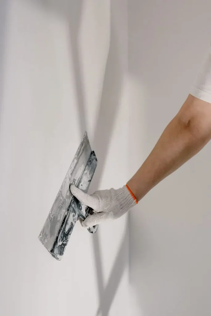 Hand met een werkhandschoen strijkt met een plamuurmes over een witte muur