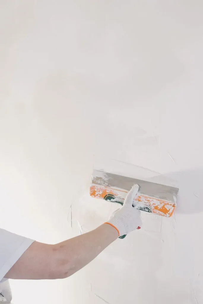 Hand met een handschoen strijkt met een spaan een witte stuclaag op een muur